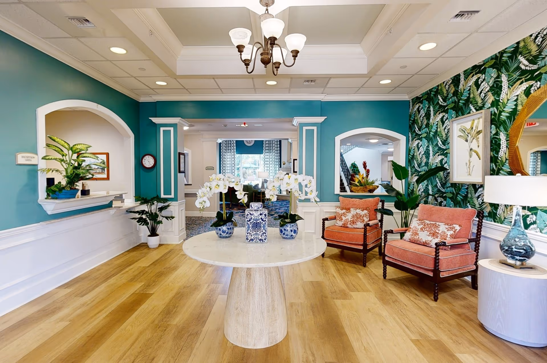 A bright and welcoming interior space with teal walls and white wainscoting. In the center, a round wooden table holds three blue and white vases with white orchids. To the right, two coral upholstered armchairs with floral cushions sit next to a small round side table with a blue and white lamp. The wall behind the chairs features green leafy patterned wallpaper, a framed botanical print, and a large round mirror. On the left, there is a reception area window with a potted plant on the ledge. The floor is light wood, and the ceiling has recessed lighting and a chandelier.