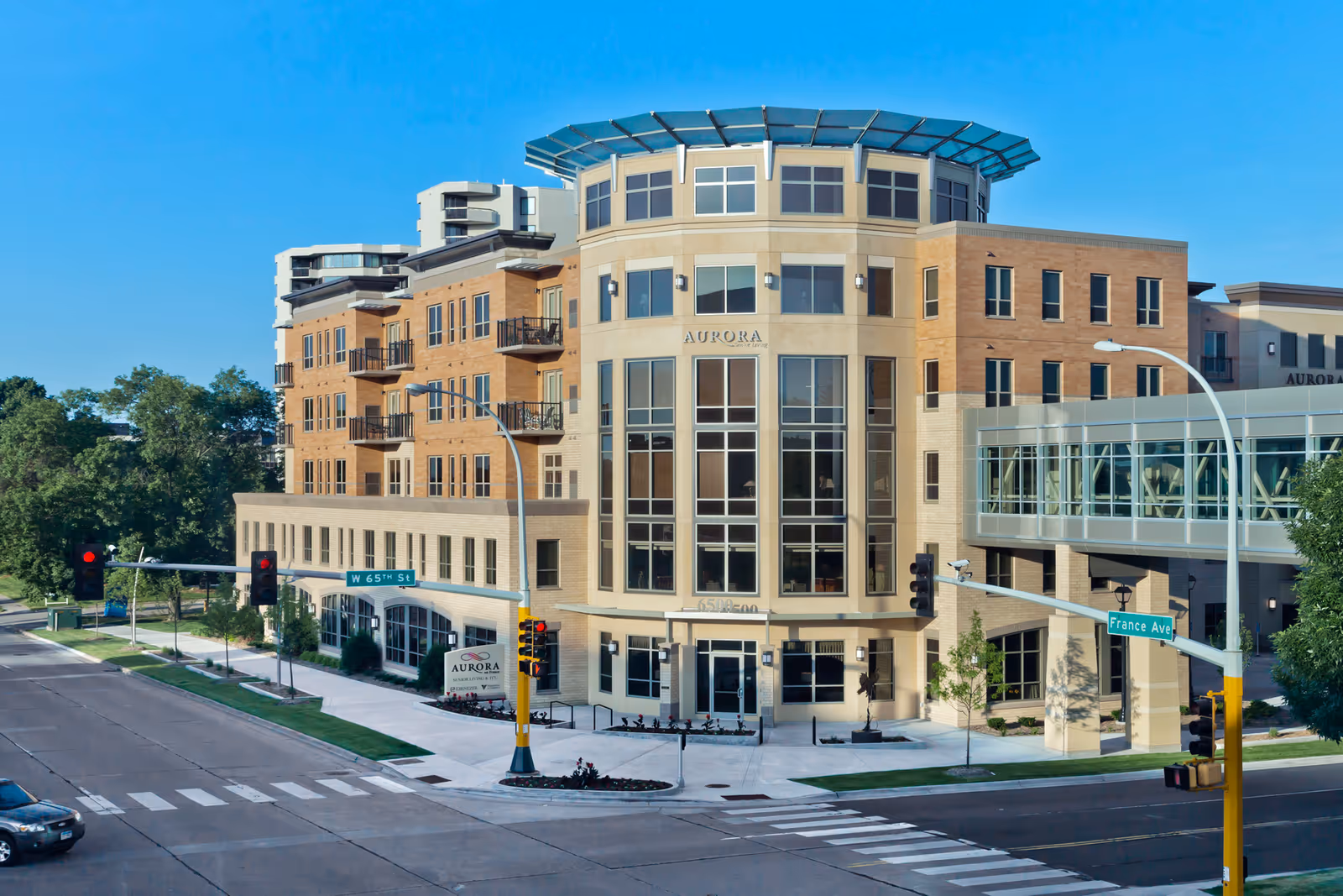 Exterior view of a multi-story senior living facility named Aurora on France, located at the corner of W 65th St and France Ave. The building features large windows, balconies, and a modern architectural design with a clear blue sky in the background.