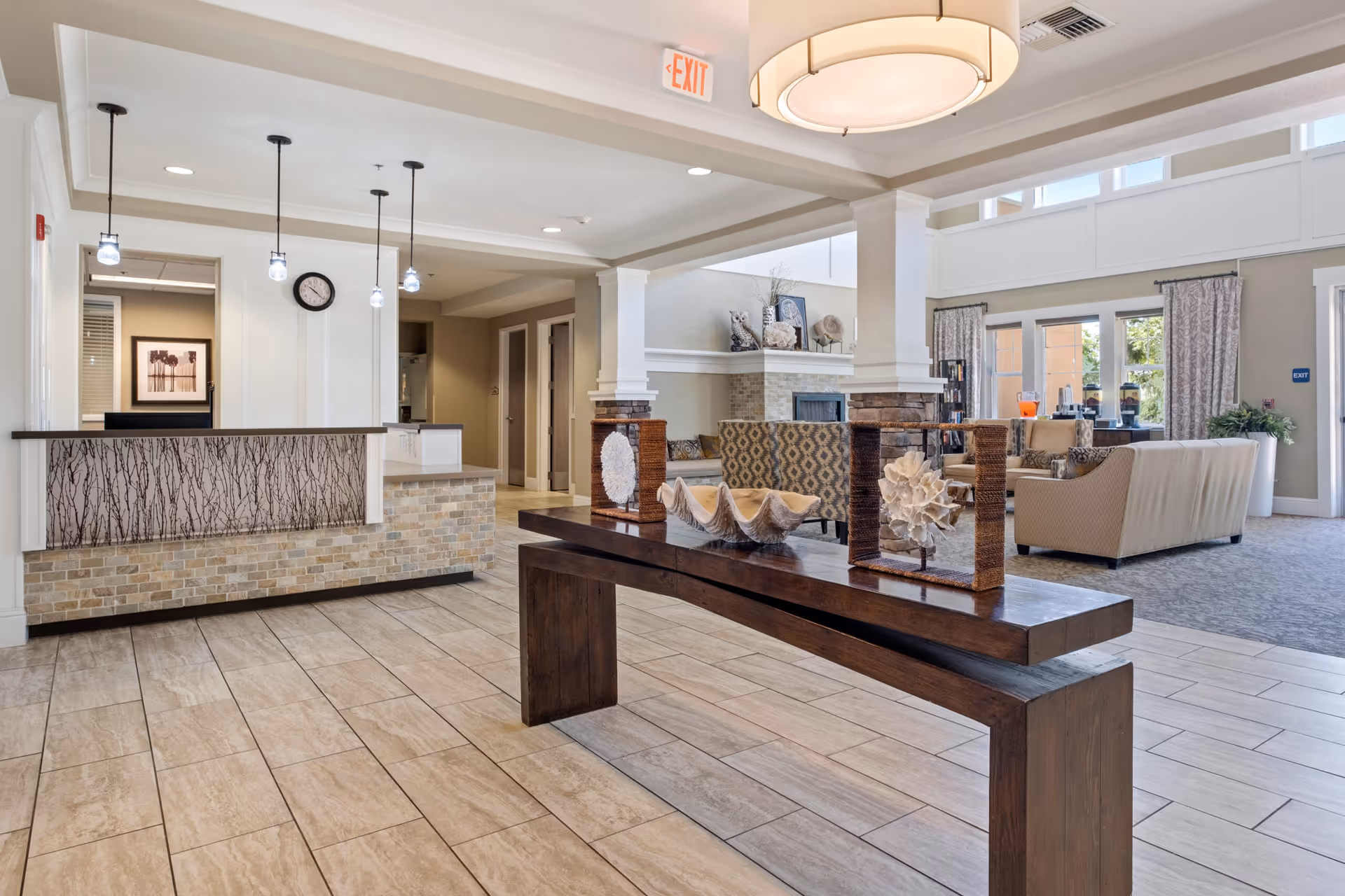 A bright and spacious senior living facility lobby with a reception desk on the left, a decorative wooden table with sea-themed ornaments in the center, and a comfortable seating area with sofas and armchairs near a fireplace in the background. Large windows with curtains allow natural light to fill the room.
