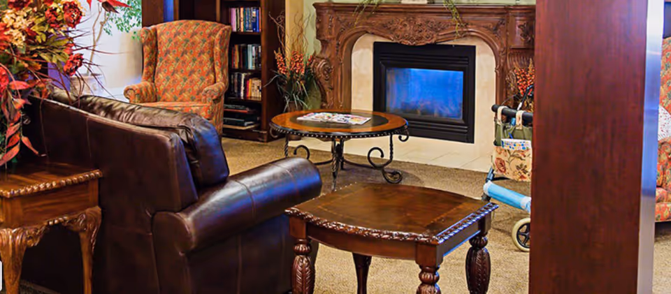 A cozy living room area with a dark brown leather sofa, a floral upholstered armchair, a wooden side table, and a round coffee table in front of an ornate wooden fireplace with a lit fire. There is a bookshelf with books and decorative plants around the room, and a walker with a floral bag is visible near the fireplace.