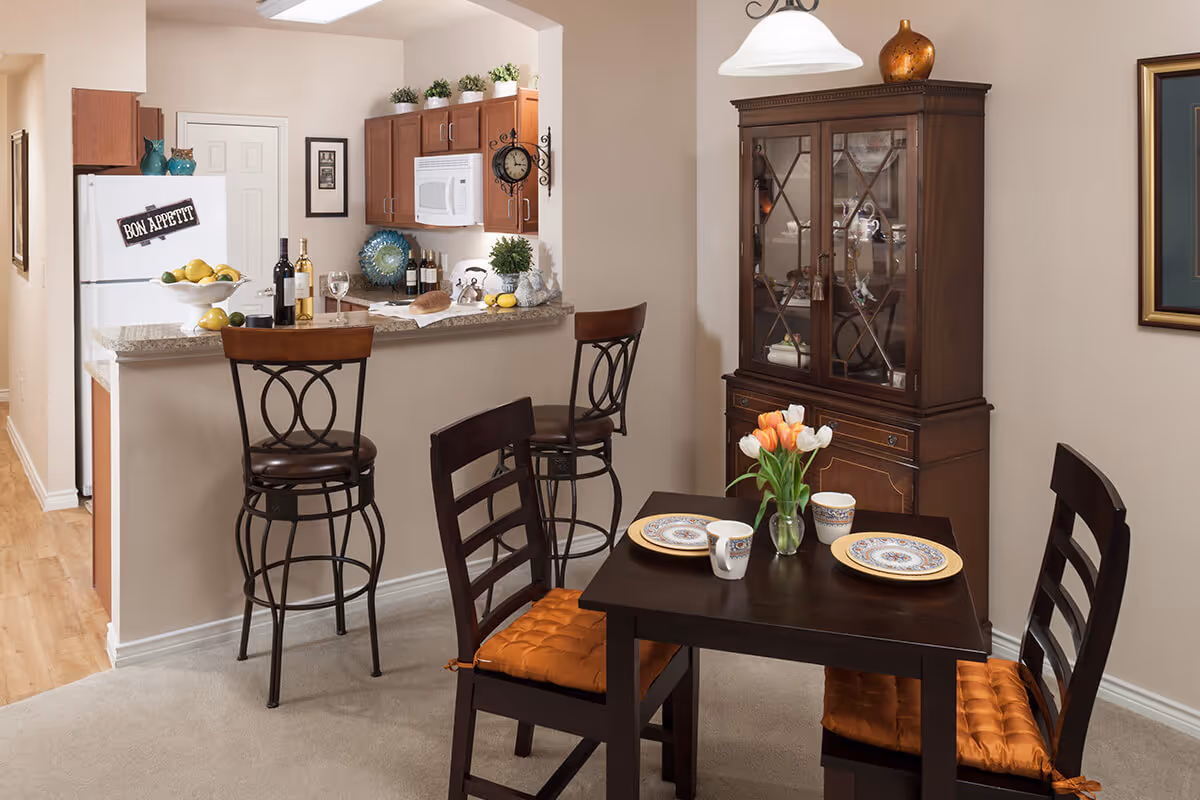 A cozy dining area with a small dark wooden table set for two, featuring plates, mugs, and a vase with orange and white tulips. Behind the table is a wooden china cabinet with glass doors displaying dishes and decorative items. To the left, there is a kitchen counter with two high chairs, a white refrigerator with a 'Bon Appetit' sign, and kitchen cabinets with various bottles and decor items.