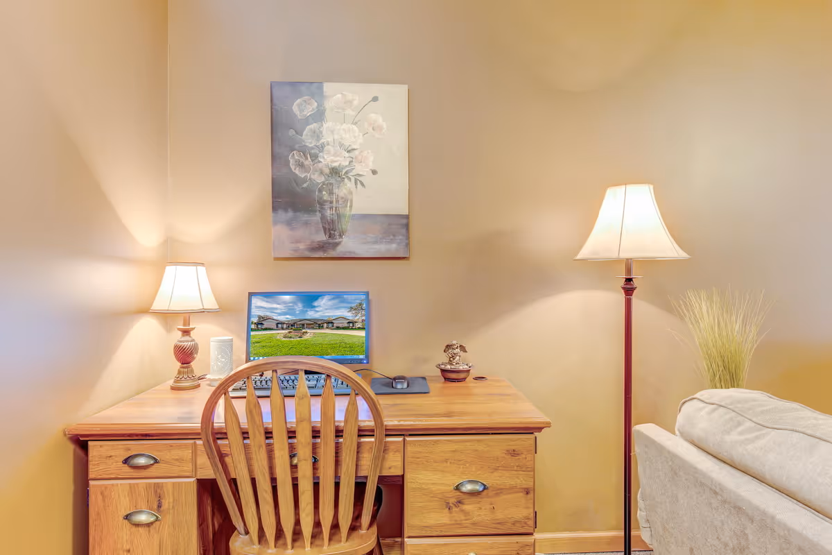 A cozy interior corner featuring a wooden desk with a matching wooden chair. On the desk, there is a table lamp, a computer monitor displaying an outdoor scene, a keyboard, a mouse, and a small decorative item. Above the desk hangs a painting of white flowers in a vase. To the right of the desk is a floor lamp and a beige sofa with a plant behind it. The walls are painted a warm beige color.