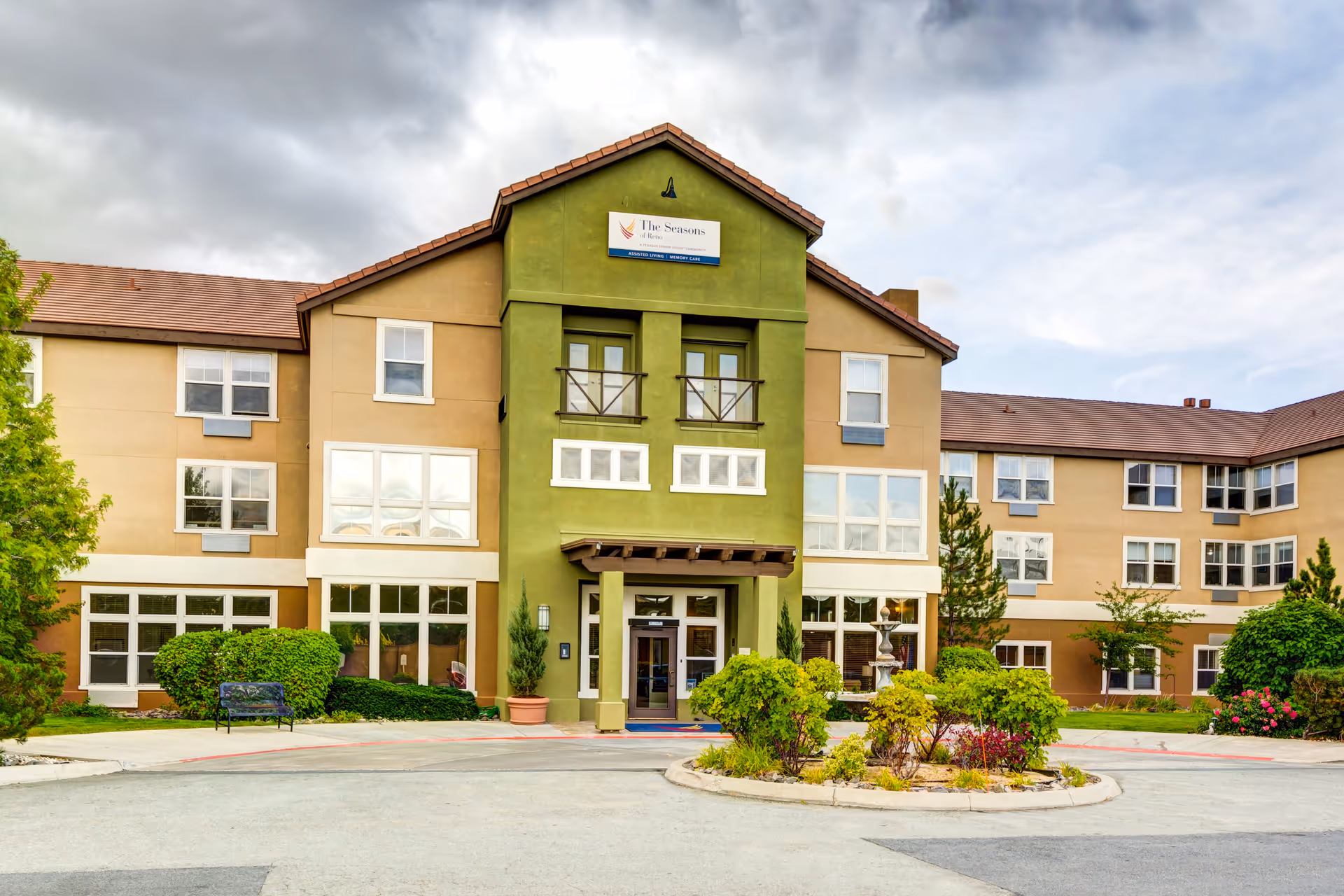Exterior front view of The Seasons of Reno senior living facility, showing a three-story building with beige and green walls, multiple windows, a central entrance with a small covered porch, and landscaped greenery including bushes and trees around the driveway.