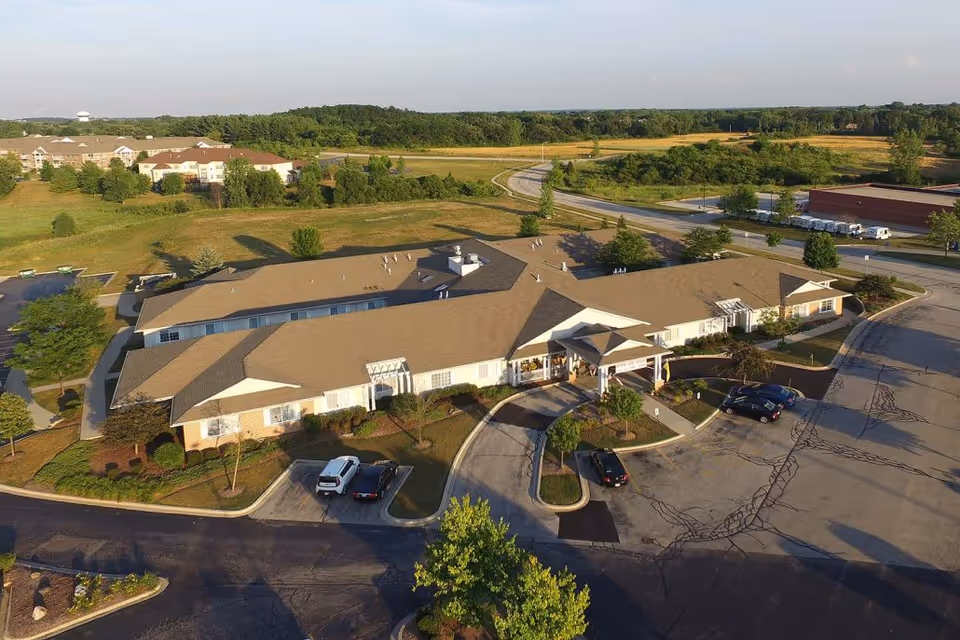 Aerial view of Brenwood Park Assisted Living facility showing a single-story building with a tan roof surrounded by parking lots, trees, and landscaped areas. The building is situated in a semi-rural area with open fields and other buildings in the distance under a clear sky.