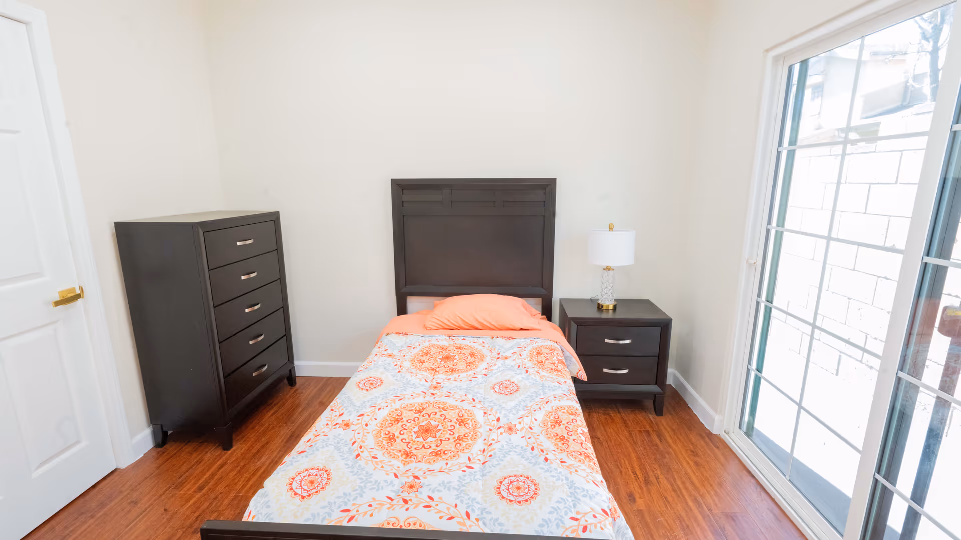 A small bedroom with a single bed featuring an orange and white patterned bedspread and an orange pillow. Next to the bed is a dark wooden nightstand with a white lamp. On the left side of the room is a matching dark wooden dresser. The room has wooden flooring, white walls, a white door, and a large window with sliding glass doors letting in natural light.
