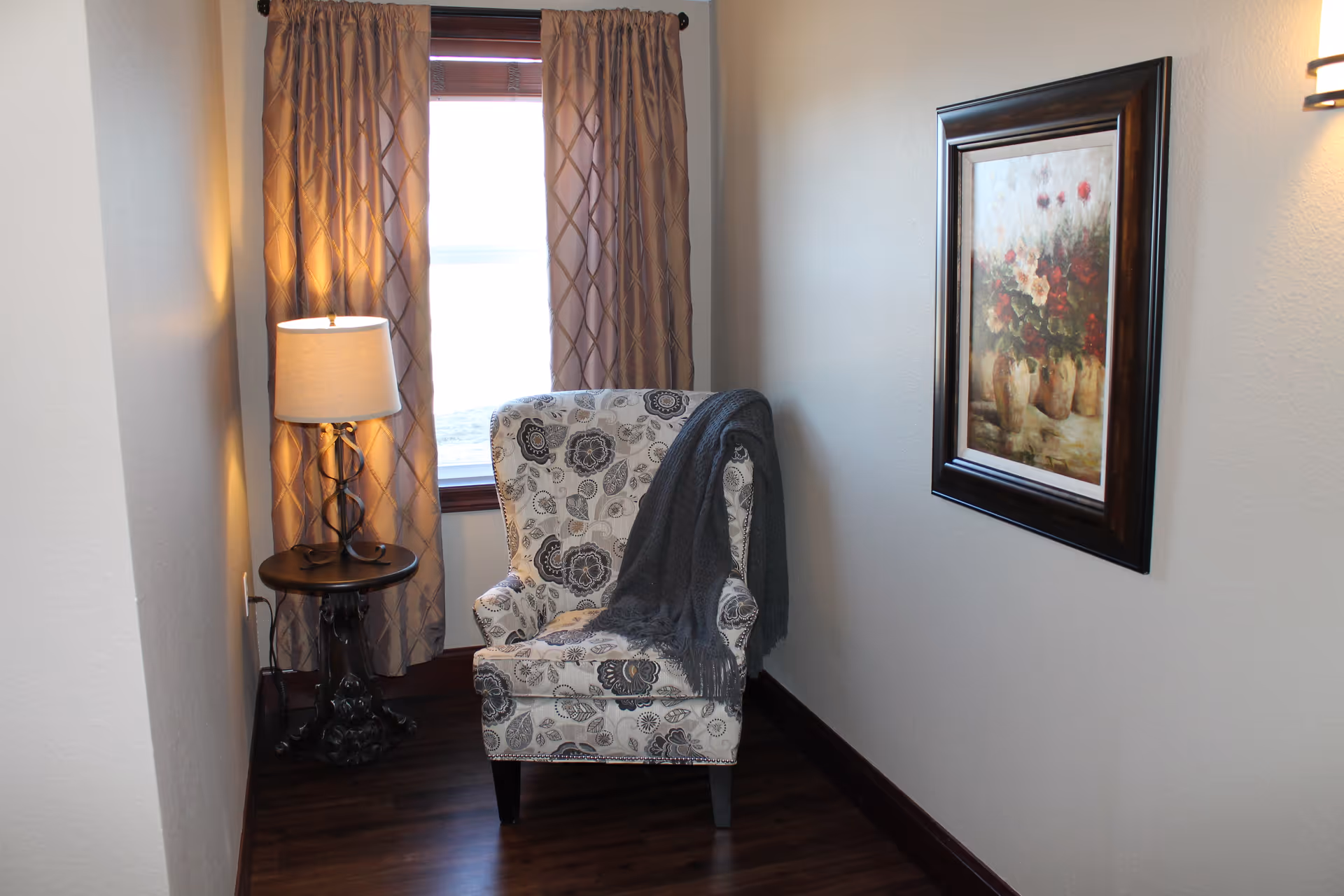 A cozy corner with a patterned armchair draped with a gray throw blanket, next to a small round table with a lamp. Behind the chair is a window with beige curtains, and a framed floral painting hangs on the adjacent wall. The floor is dark wood.