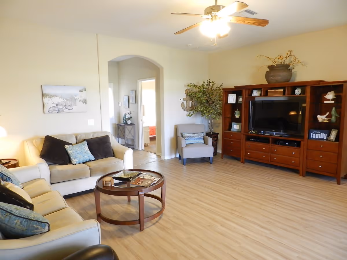 A cozy living room with light-colored sofas adorned with blue and black cushions, a round wooden coffee table with magazines, a single armchair with a striped cushion, a large wooden entertainment center with a flat-screen TV and decorative items, a potted plant, and a ceiling fan with lights. The room has light wood flooring and beige walls, with an arched doorway leading to another room.