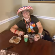 An elderly woman wearing a colorful sombrero and glasses is sitting at a wooden table with a plate of food and two cups in front of her. She is smiling and appears to be enjoying herself in an indoor setting with beige walls and white trim.