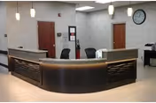 Reception area with a curved dark wood desk, two office chairs behind the desk, two closed wooden doors on a light gray wall, a clock on the wall, and ceiling lights above.