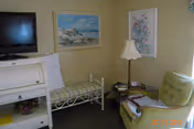 A cozy living room area with a green upholstered armchair, a white metal bench with a cushion, a white roll-top desk, a floor lamp, and two framed pictures on the walls. A flat-screen TV is mounted on the wall above the desk.