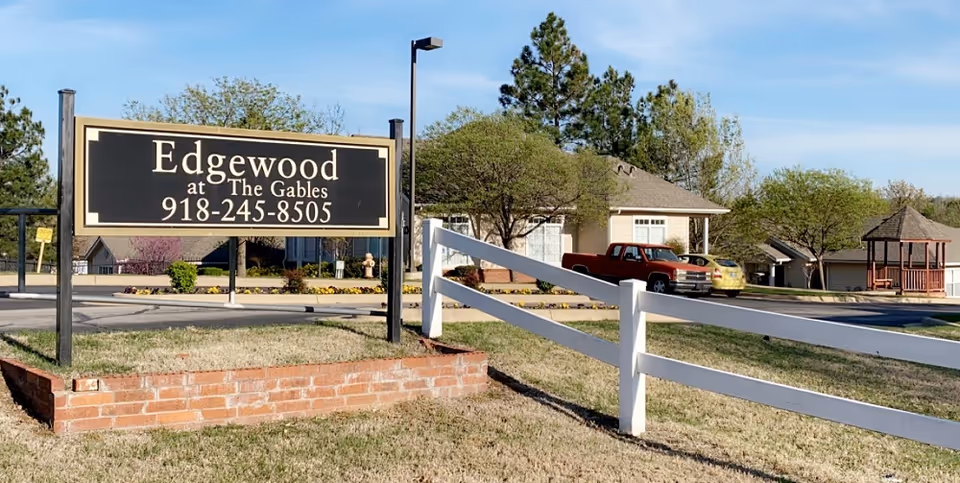 Outdoor view of the Edgewood at The Gables senior living facility sign with phone number 918-245-8505. The sign is mounted on black posts above a low brick wall. Behind the sign, there is a driveway, a beige building, parked vehicles, trees, and a gazebo under a clear blue sky.