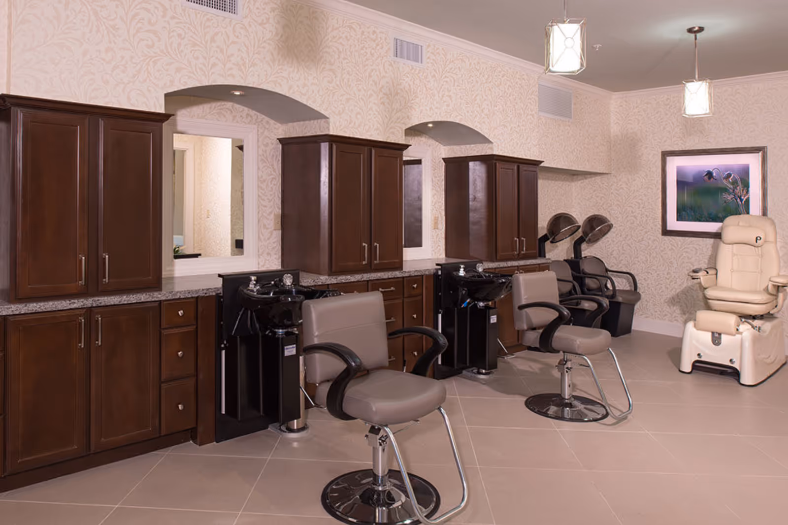 Interior view of a salon area in a retirement community featuring two salon chairs in front of sinks and mirrors, two hair dryer chairs in the background, and a beige pedicure chair with a framed floral picture on the wall.