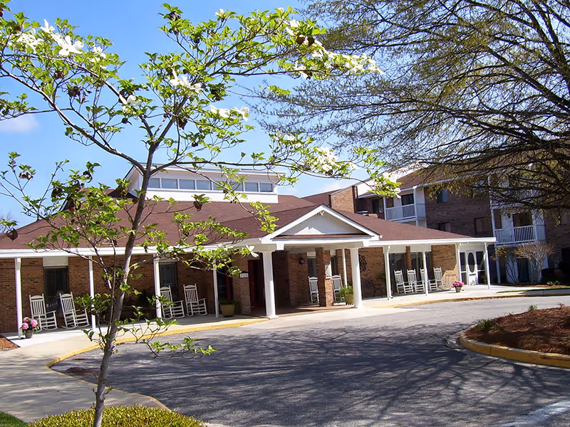 Exterior view of a brick retirement community building with a covered entrance supported by white columns. Several white rocking chairs are placed along the porch. Trees with green leaves and white flowers are visible in the foreground under a clear blue sky.