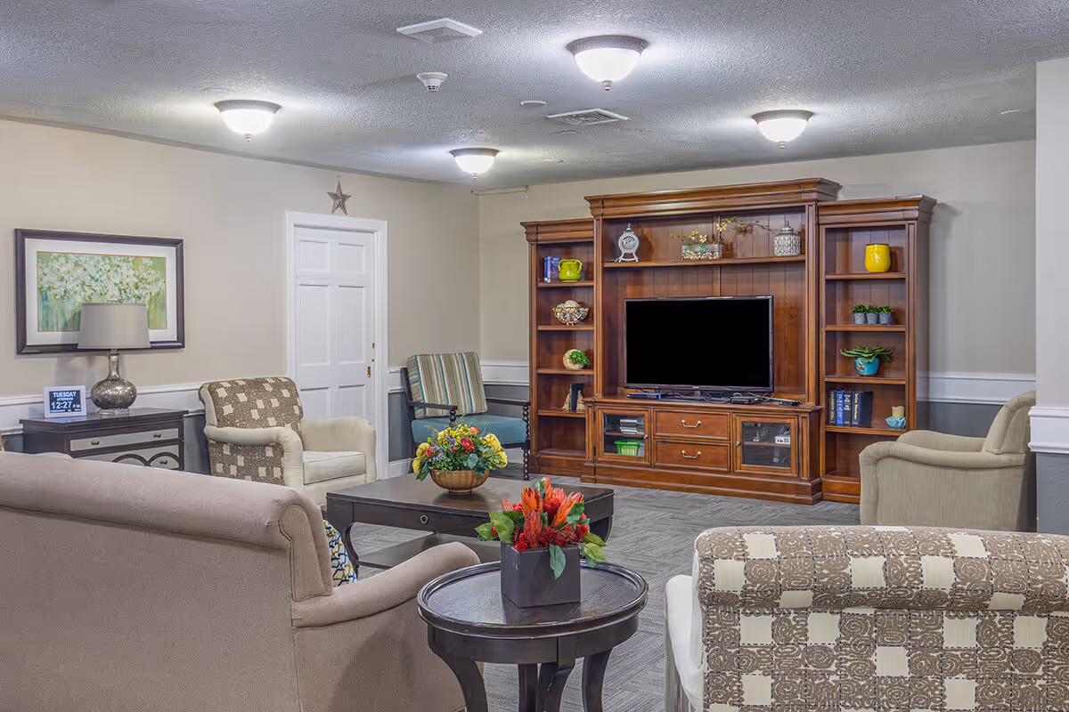 A cozy senior living room with multiple upholstered armchairs and sofas arranged around a dark wooden coffee table and side table with flower arrangements. A large wooden entertainment center with a flat-screen TV and decorative items is against the wall. The room has soft lighting from ceiling fixtures and neutral-colored walls with a framed floral painting and a digital clock on a side table.