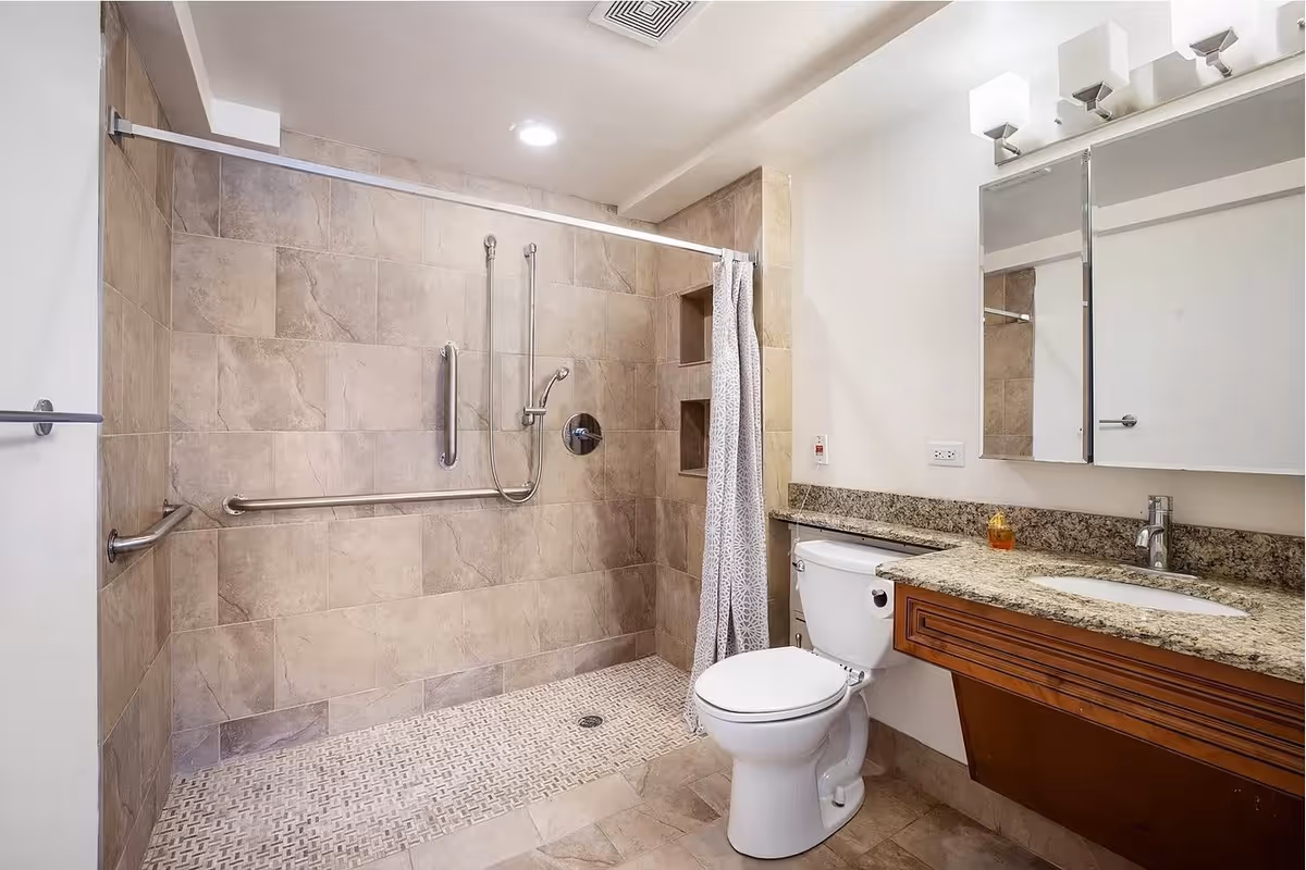 A modern bathroom with beige tiled walls and floor. The walk-in shower area has a handheld showerhead, grab bars, and a shower curtain. There is a white toilet next to a granite countertop with a built-in sink and a wooden vanity. Above the sink is a large mirror with three light fixtures.
