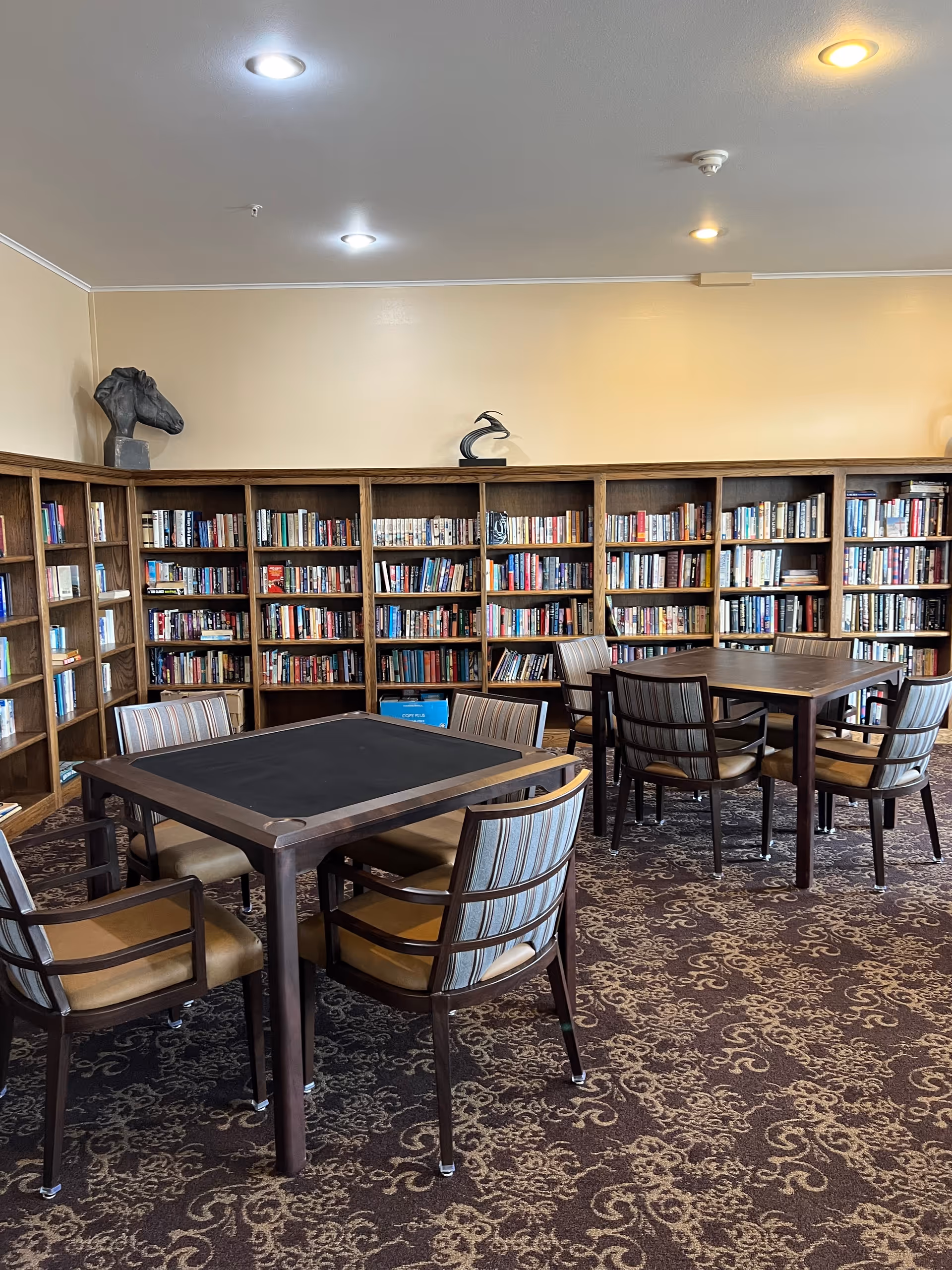 A cozy library room with wooden bookshelves filled with books lining the walls. Two square tables with four cushioned chairs each are placed on a patterned carpet. Decorative sculptures are displayed on top of the bookshelves under ceiling lights.