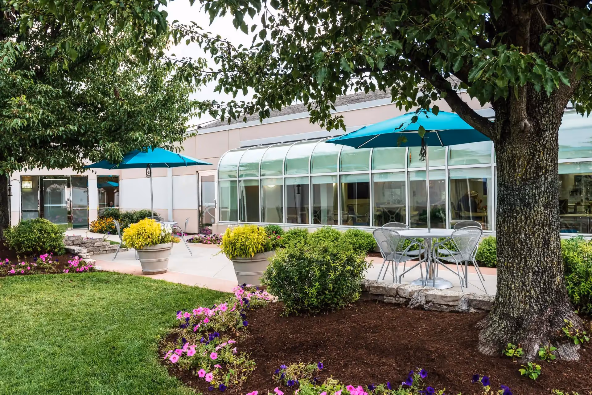 Outdoor patio area at Delmar Gardens South with metal tables and chairs under teal umbrellas, surrounded by green bushes, colorful flowers, and trees. The building features large windows and a glass-covered walkway.