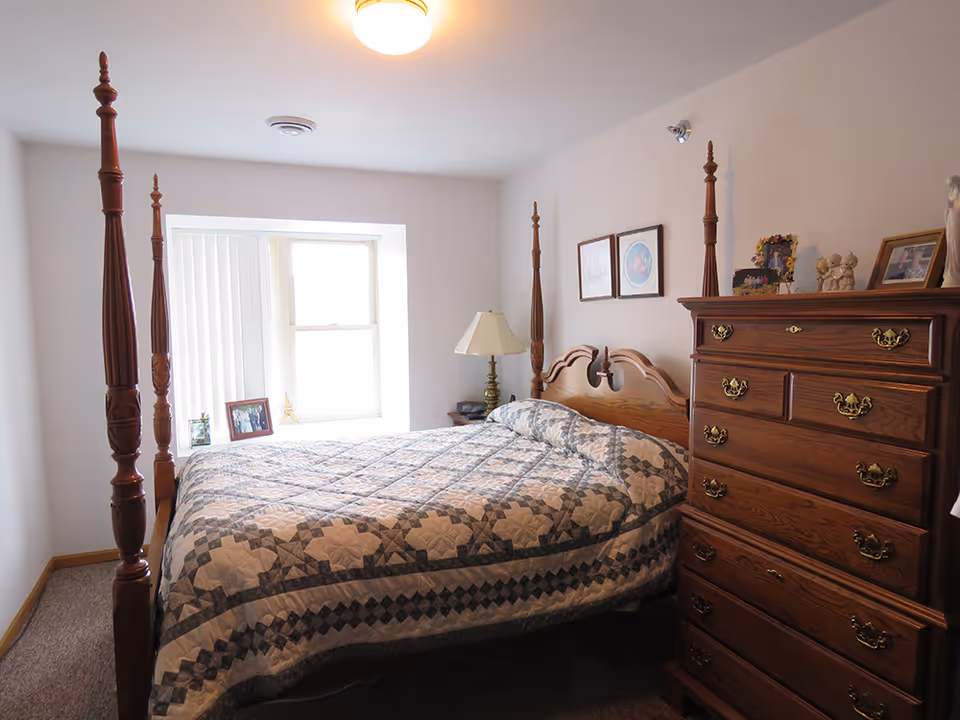 A cozy bedroom with a wooden four-poster bed covered with a patterned quilt. There is a wooden dresser with brass handles on the right side, a bedside table with a lamp, and framed pictures on the wall and dresser. A window with vertical blinds lets in natural light.