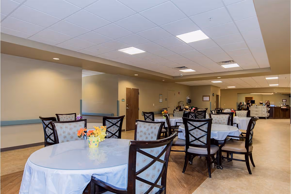 Interior view of a dining room in a senior living facility with round tables covered with white tablecloths and decorated with small flower arrangements. Several chairs with patterned upholstery surround the tables. The room has a beige color scheme, a drop ceiling with fluorescent lighting, and a reception desk area in the background.