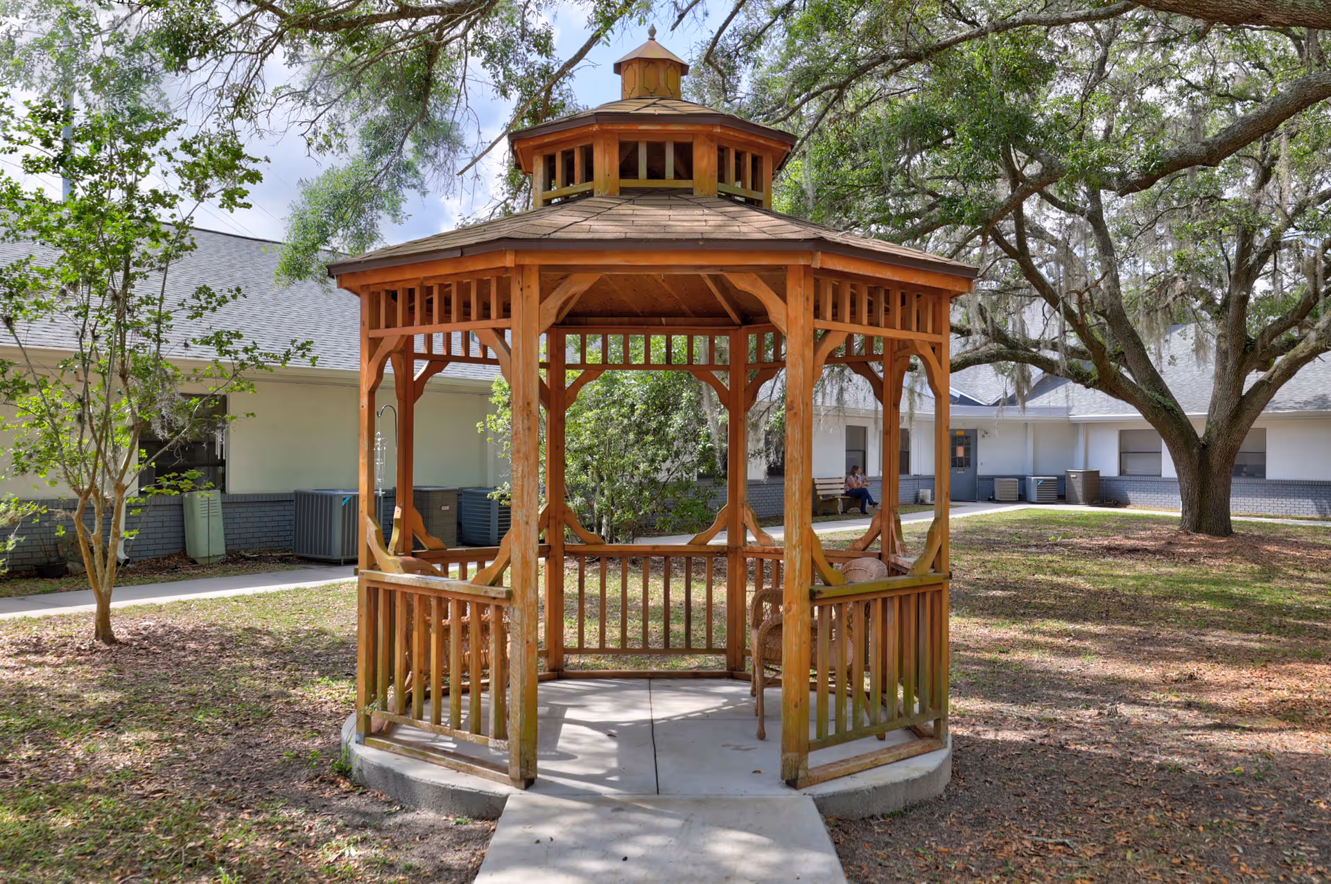 A wooden gazebo with a shingled roof situated on a concrete pad in a grassy courtyard area. Surrounding the gazebo are trees and bushes, with a building featuring white walls and a gray roof in the background. A person is sitting on a bench near the building.