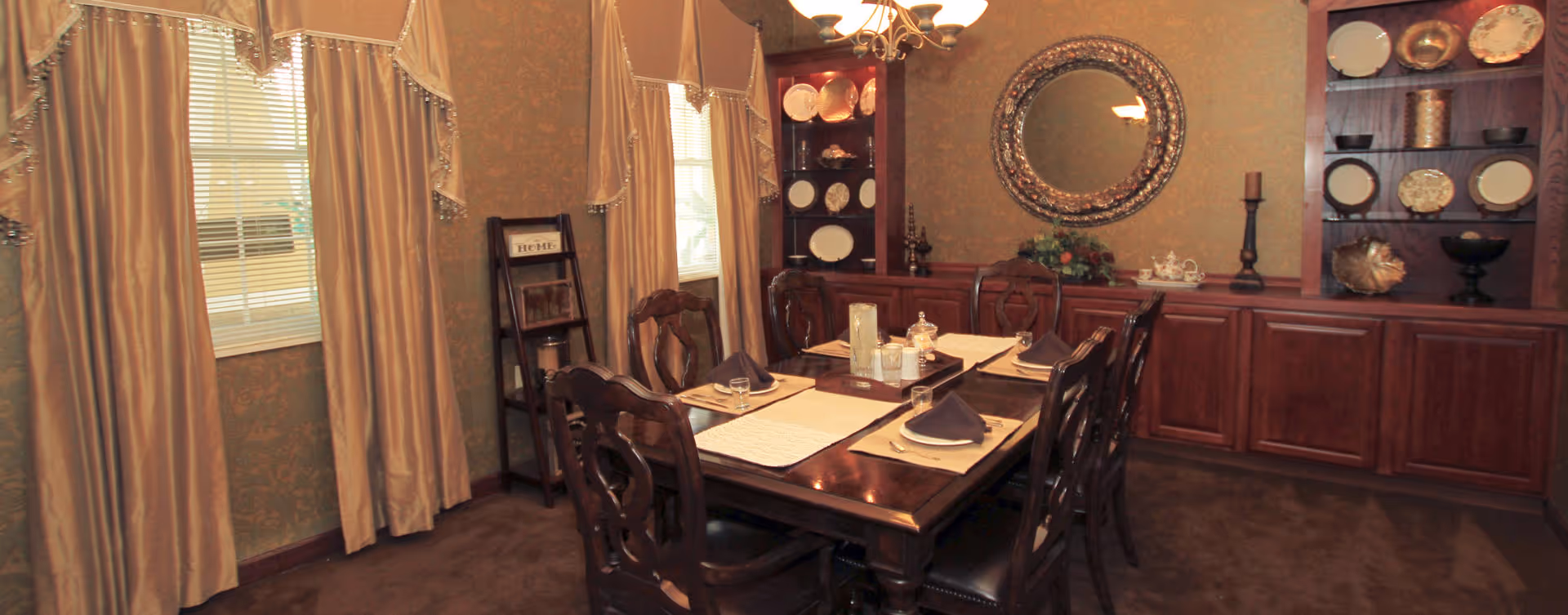 A formal dining room with a dark wooden table set for six people, featuring placemats, napkins, and glassware. The room has beige curtains on the windows, a chandelier overhead, and built-in wooden cabinets with decorative plates and items. A large round mirror hangs on the wall above the cabinets.
