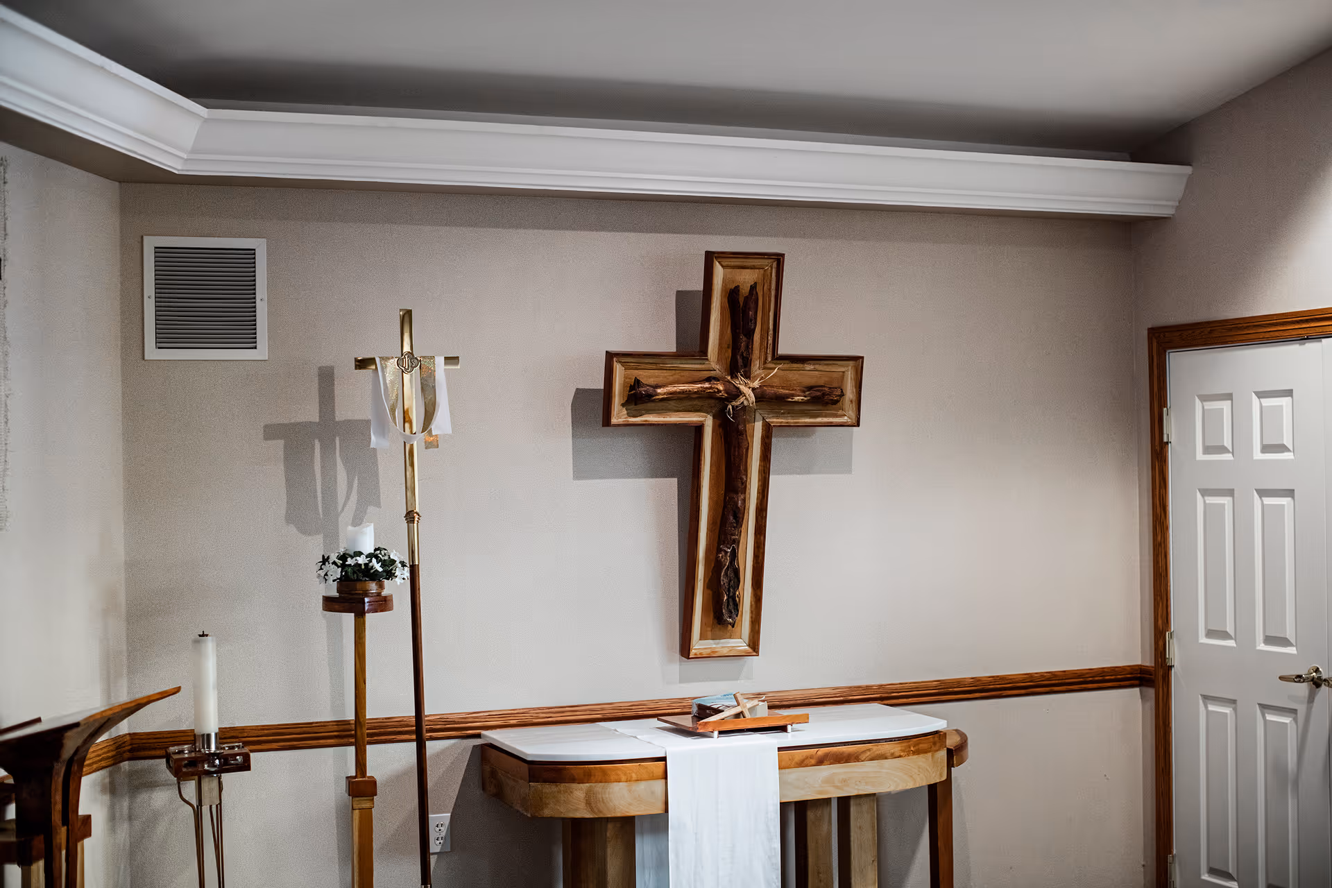 Interior room with a wooden cross mounted on the wall above a small wooden table draped with a white cloth. To the left, there is a tall wooden stand holding a candle and a floral arrangement, and a white candle on a metal holder. The room has beige walls with wooden trim and a white door on the right side.