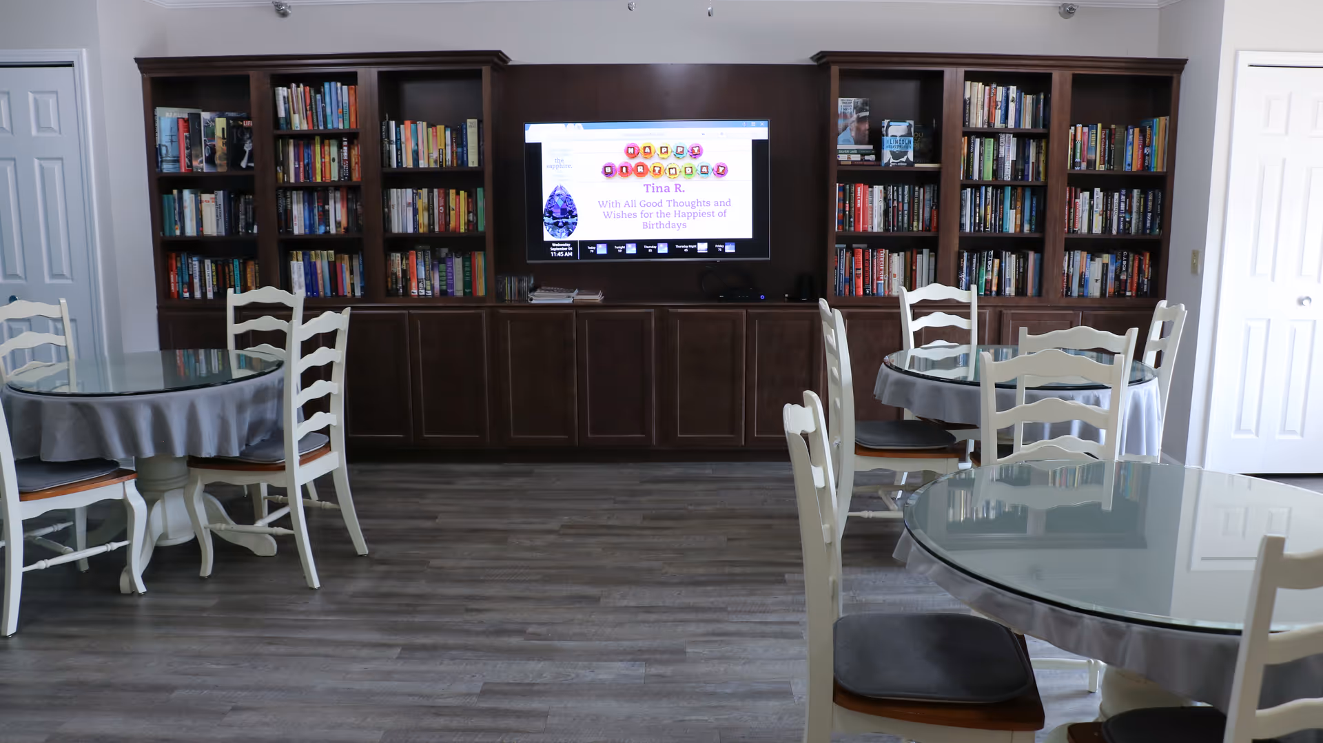 Communal room with round glass-topped tables and chairs facing built-in bookshelves and a wall-mounted TV.