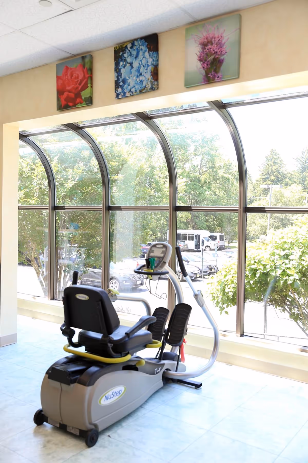 An indoor fitness room with a NuStep exercise bike near large curved windows and floral wall art.