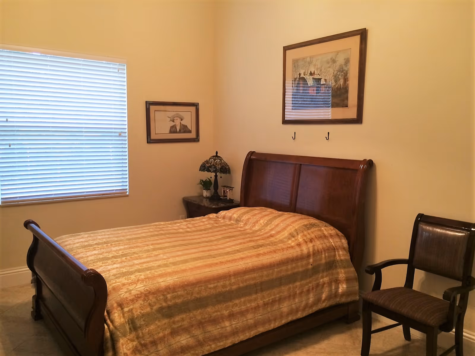 A tidy bedroom with a wooden sleigh bed covered by a patterned bedspread, a nightstand with a lamp, framed pictures on the walls, a window with blinds, and a chair.