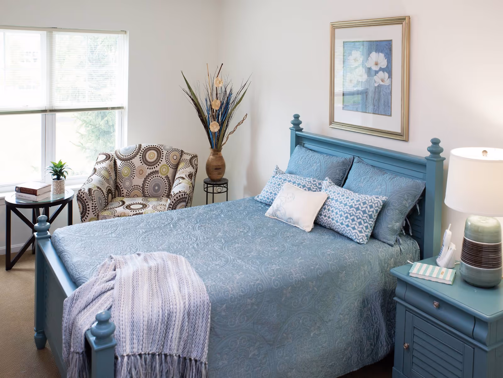 A cozy bedroom with a blue wooden bed frame and matching blue bedding, including multiple pillows. A light blue throw blanket is draped over the foot of the bed. To the right of the bed is a blue nightstand with a lamp, a phone, and a small book. To the left, there is a patterned armchair next to a round side table with two books and a small plant. A large window with blinds lets in natural light, and a framed floral artwork hangs on the wall above the bed.