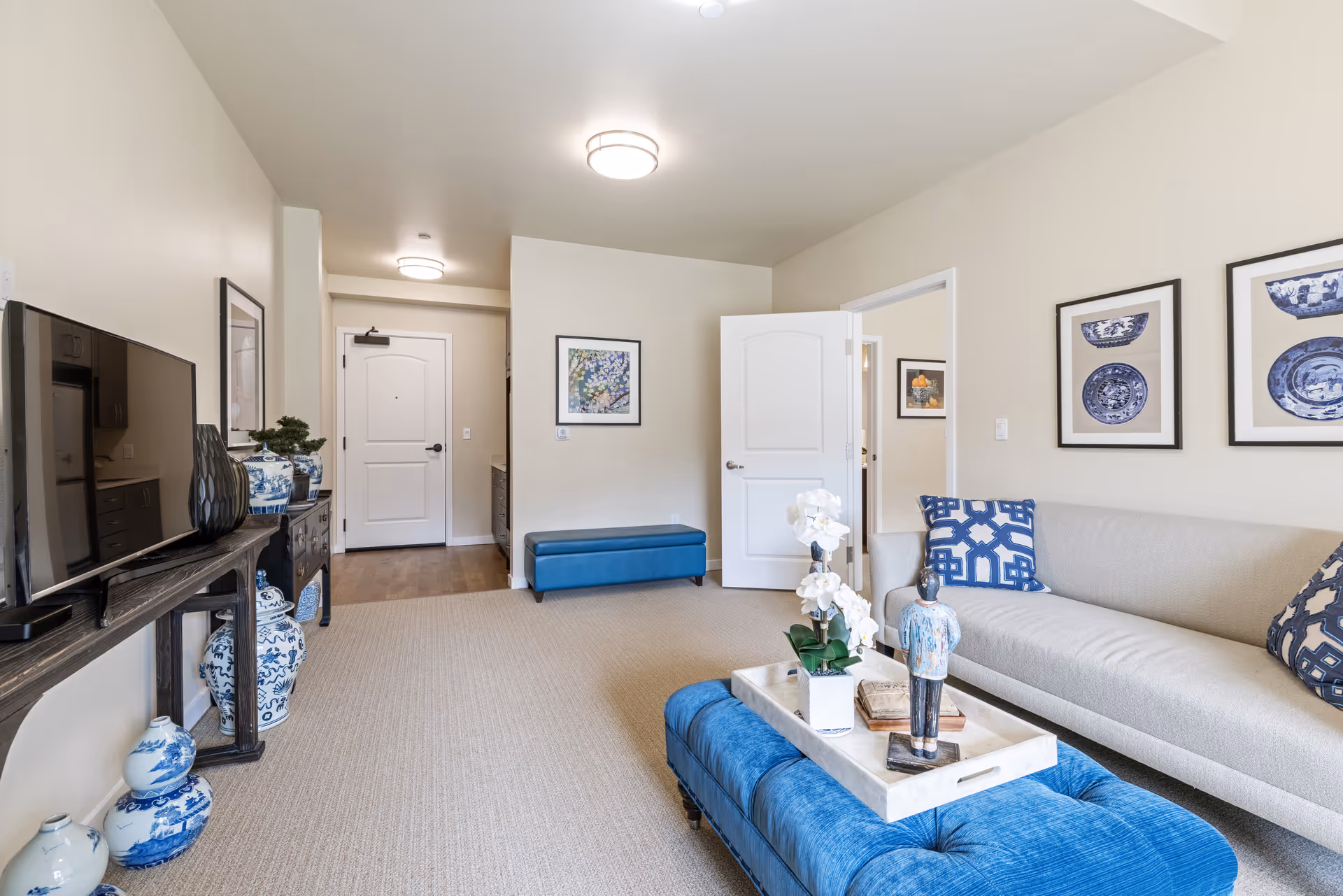 Bright living room with a beige sofa, blue tufted ottoman coffee table, TV on a console, and an entry door in the background.