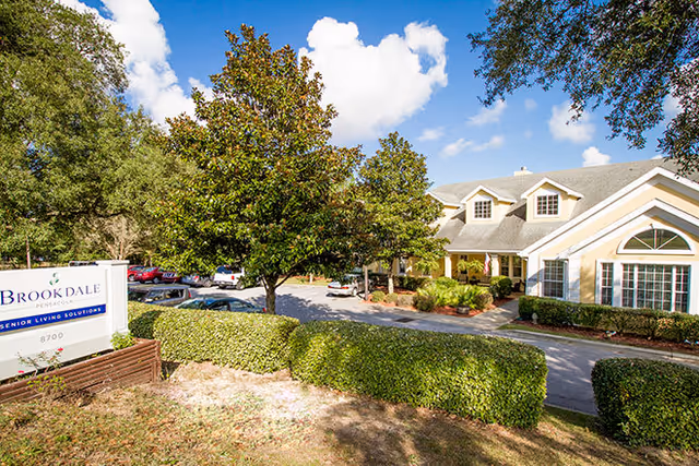 Front exterior of Brookdale Pensacola senior living facility showing the entrance, landscaped hedges, parking lot and a sign.