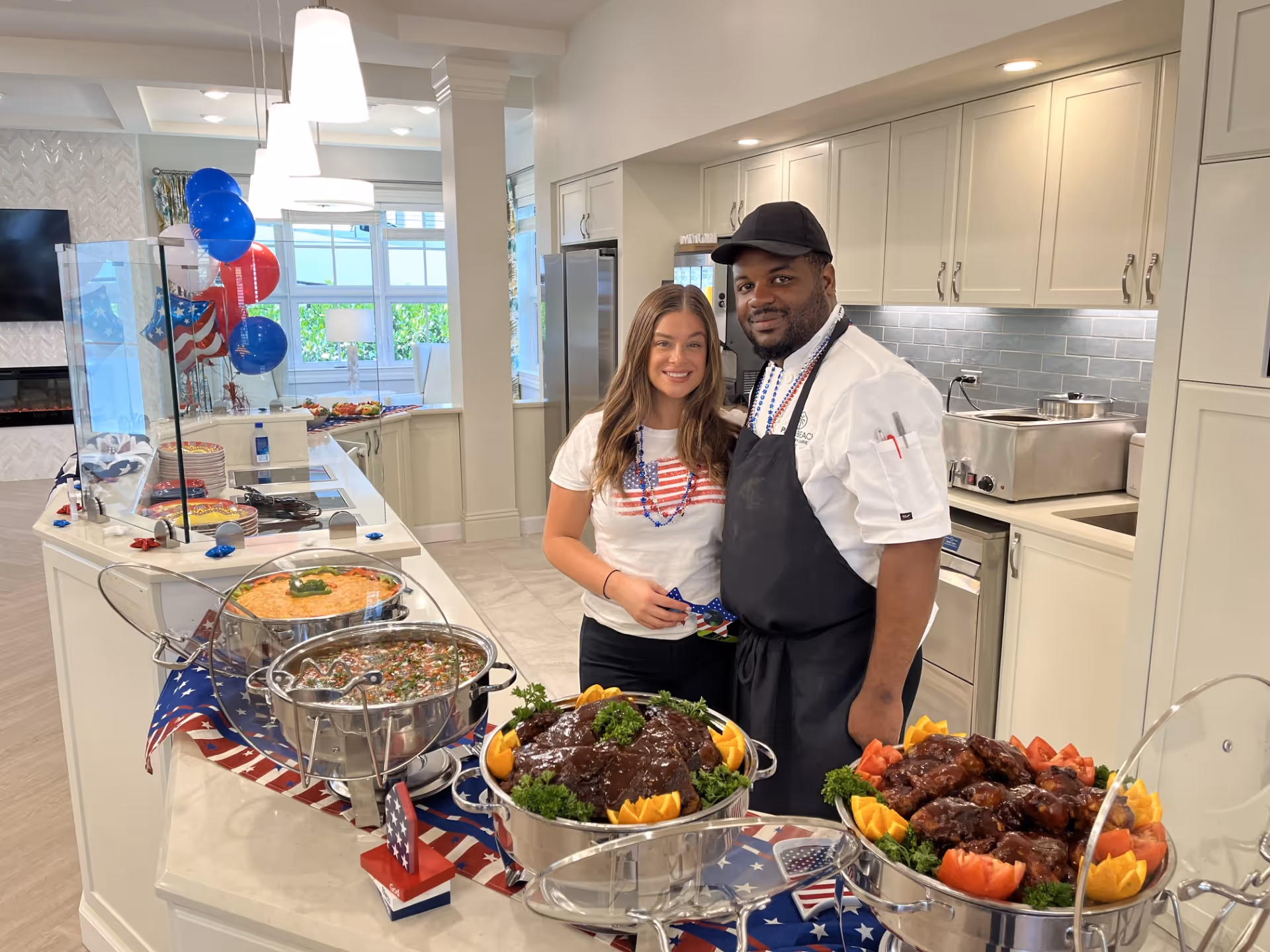 A smiling woman and a chef stand behind a buffet of chafing dishes in a decorated kitchen with American-themed balloons and food displays.