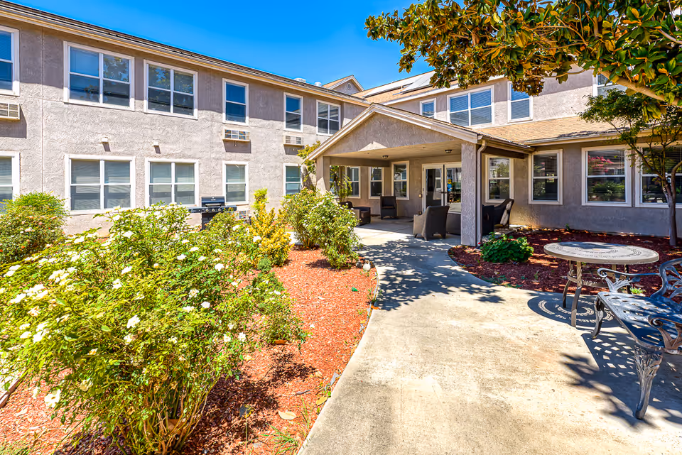 Outdoor courtyard and covered entrance of a senior living building with a walkway, seating, and landscaped flower beds.