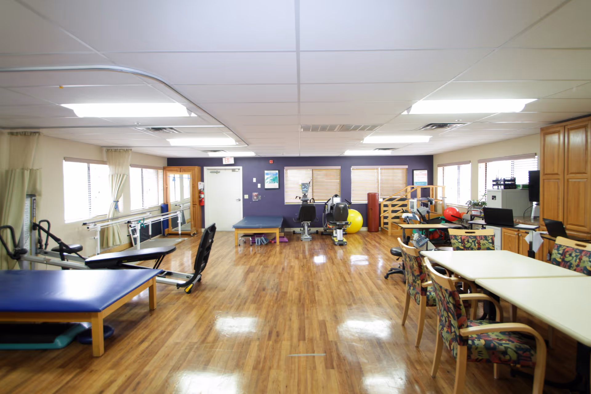 A spacious room with wooden flooring and white ceiling lights, featuring exercise equipment such as stationary bikes, parallel bars, therapy tables, and various physical therapy tools. There are tables and chairs with floral upholstery on the right side, and large windows with beige curtains letting in natural light.