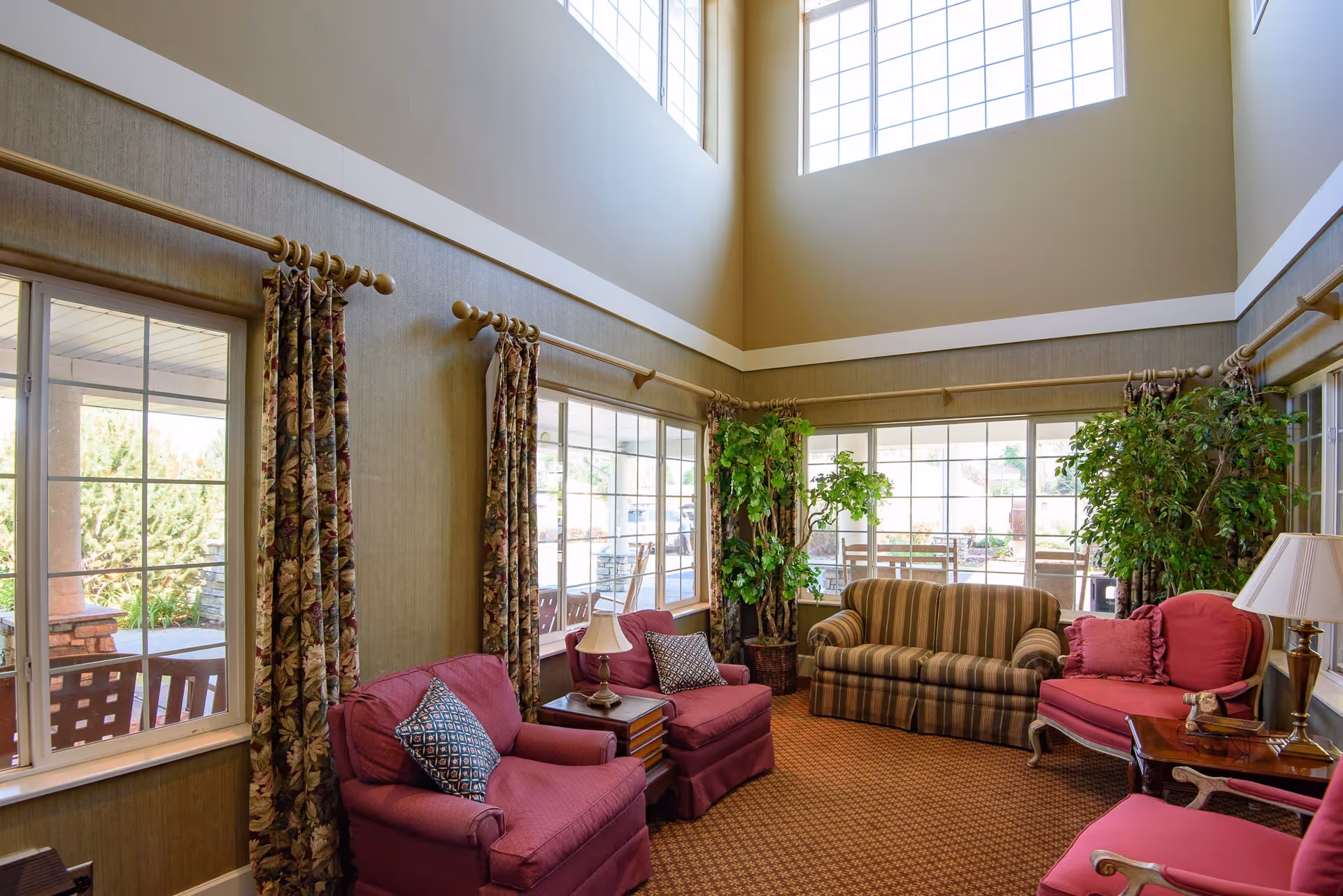 Sunlit two-story lounge with large windows, striped sofa, upholstered armchairs, side tables and potted plants.