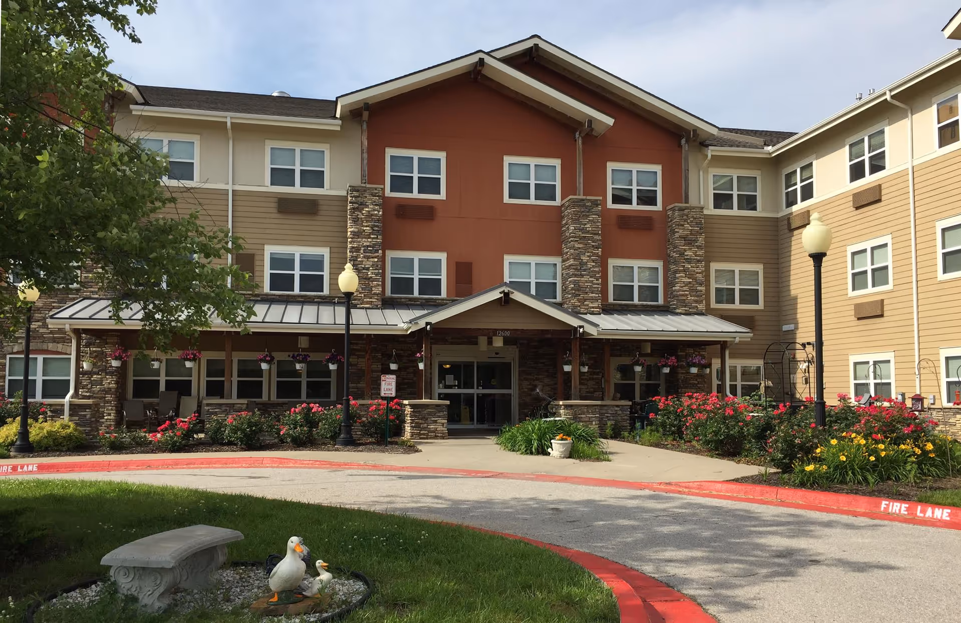 Front exterior view of Delaware Highlands Assisted Living facility showing a three-story building with beige and brown siding, stone accents, multiple windows, a covered entrance, flower beds with pink and yellow flowers, two street lamps, a small garden area with a stone bench and duck statues, and a curved driveway marked with red fire lane curbs.
