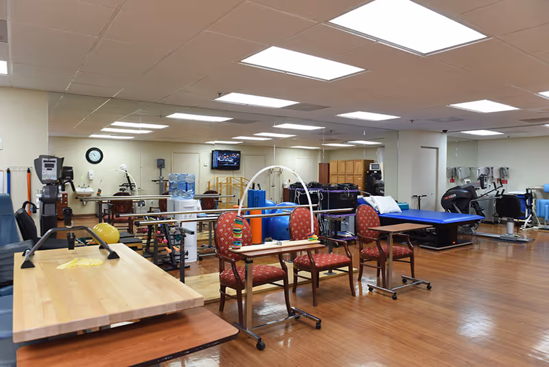 Interior view of a rehabilitation and physical therapy room with exercise equipment, parallel bars, chairs with attached activity boards, a treatment table, and a water cooler. The room has wooden flooring, a mirrored wall, and ceiling lights.