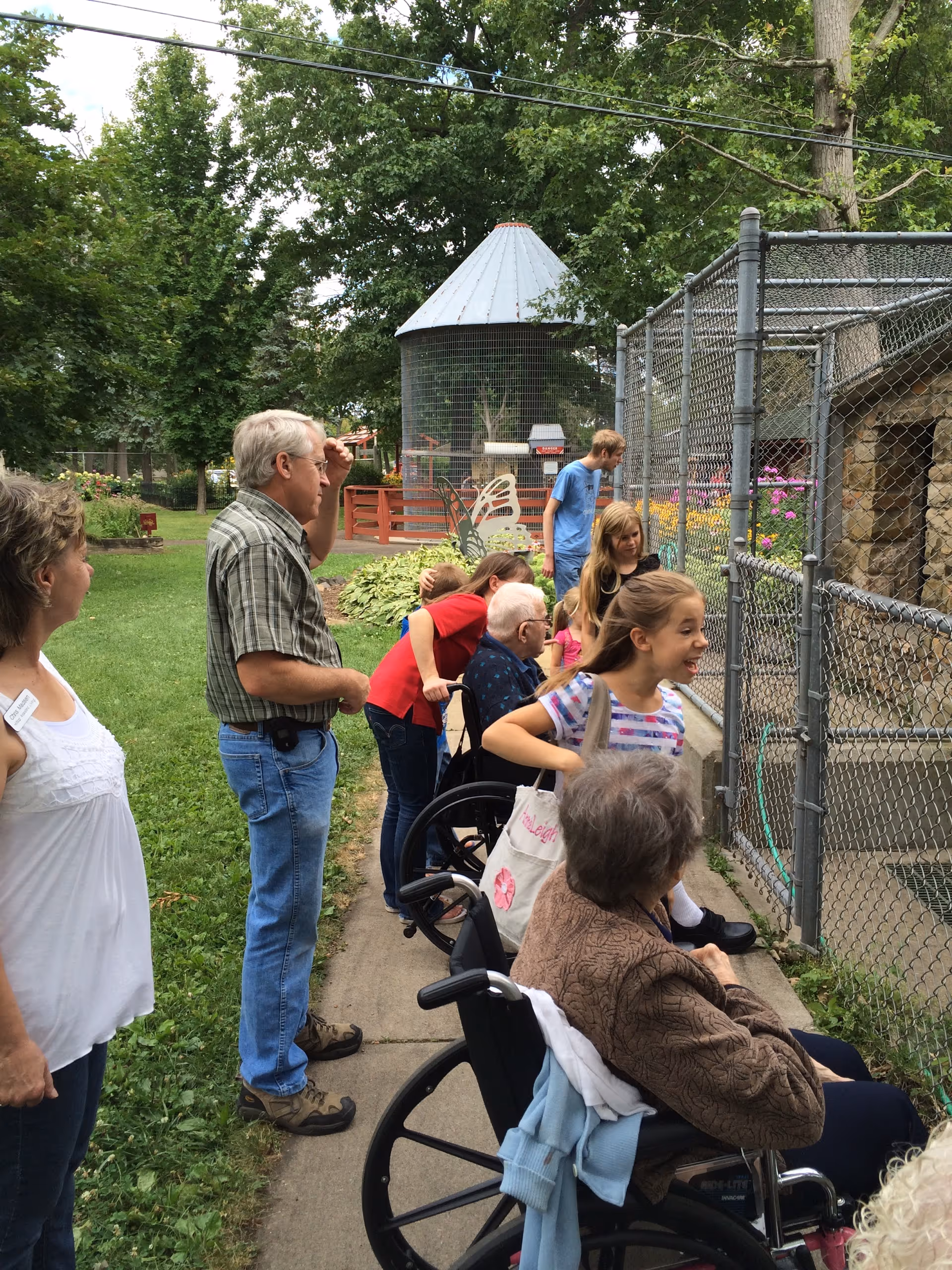 A group of people, including elderly individuals in wheelchairs and children, are gathered outside near a fenced enclosure with greenery and trees in the background. They appear to be observing something inside the enclosure on a sunny day.