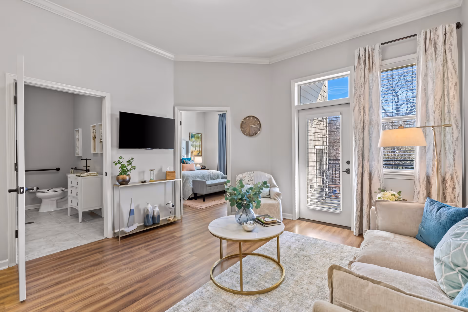 A bright living room with a beige sofa adorned with blue and patterned pillows, a round coffee table with a vase of greenery and books, and a floor lamp. There is a door with glass panels and windows with curtains letting in natural light. Adjacent rooms include a bathroom with a toilet and vanity, and a bedroom with a bed and nightstand. The floor is wooden and the walls are painted light gray.
