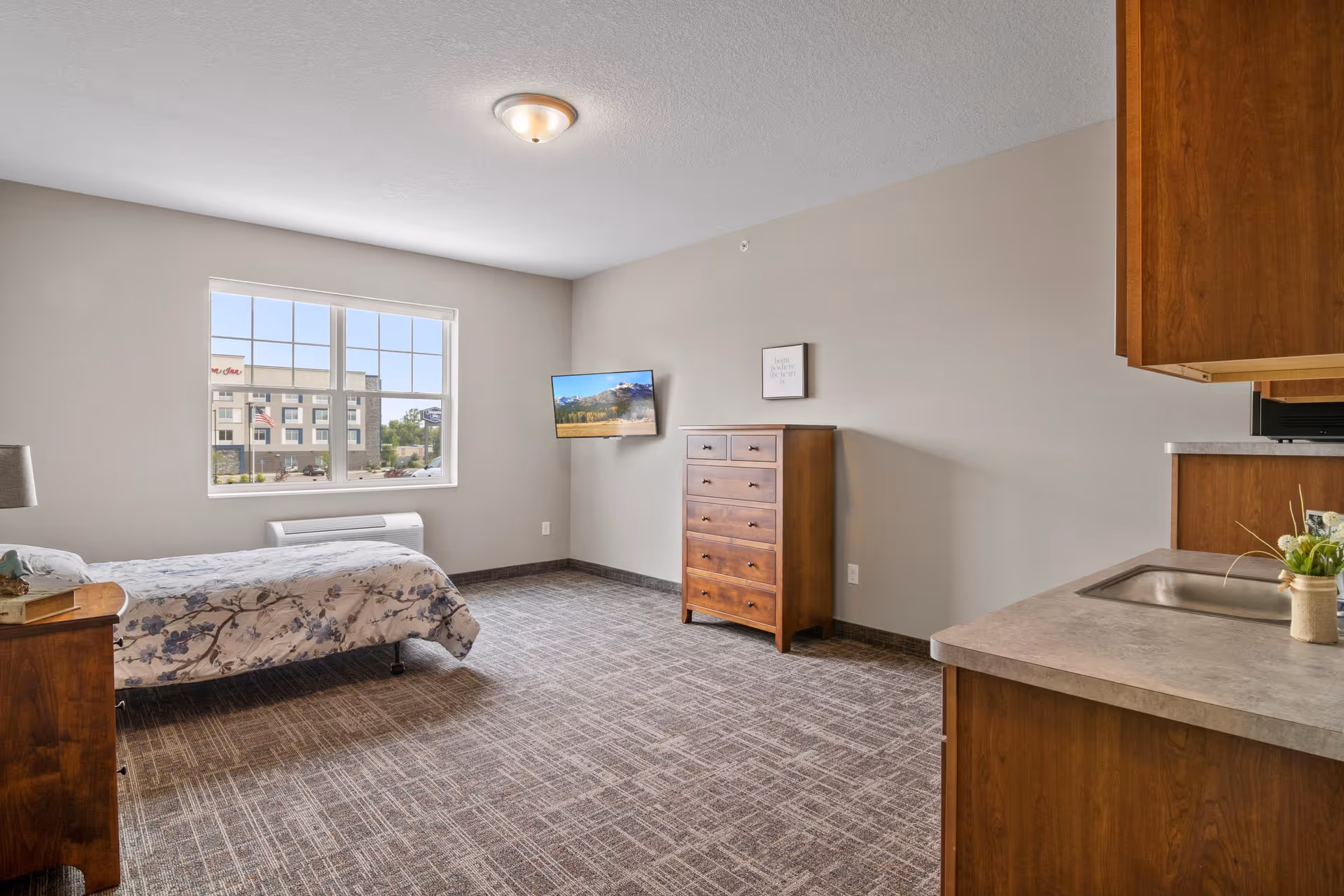 A bright studio bedroom in a senior living facility with a bed, dresser, wall-mounted TV, and a small kitchenette area.