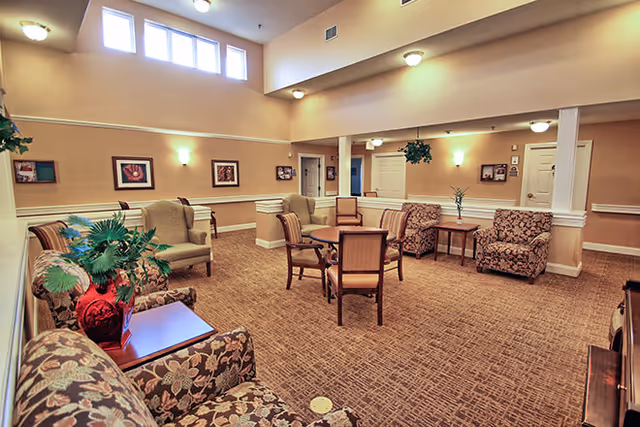 A spacious and well-lit common area with beige walls and carpeted floor. The room features multiple seating arrangements including floral and solid upholstered armchairs, a round wooden table with four chairs, and side tables with decorative plants. High windows near the ceiling allow natural light to enter, and framed artwork decorates the walls. Ceiling lights provide additional illumination.