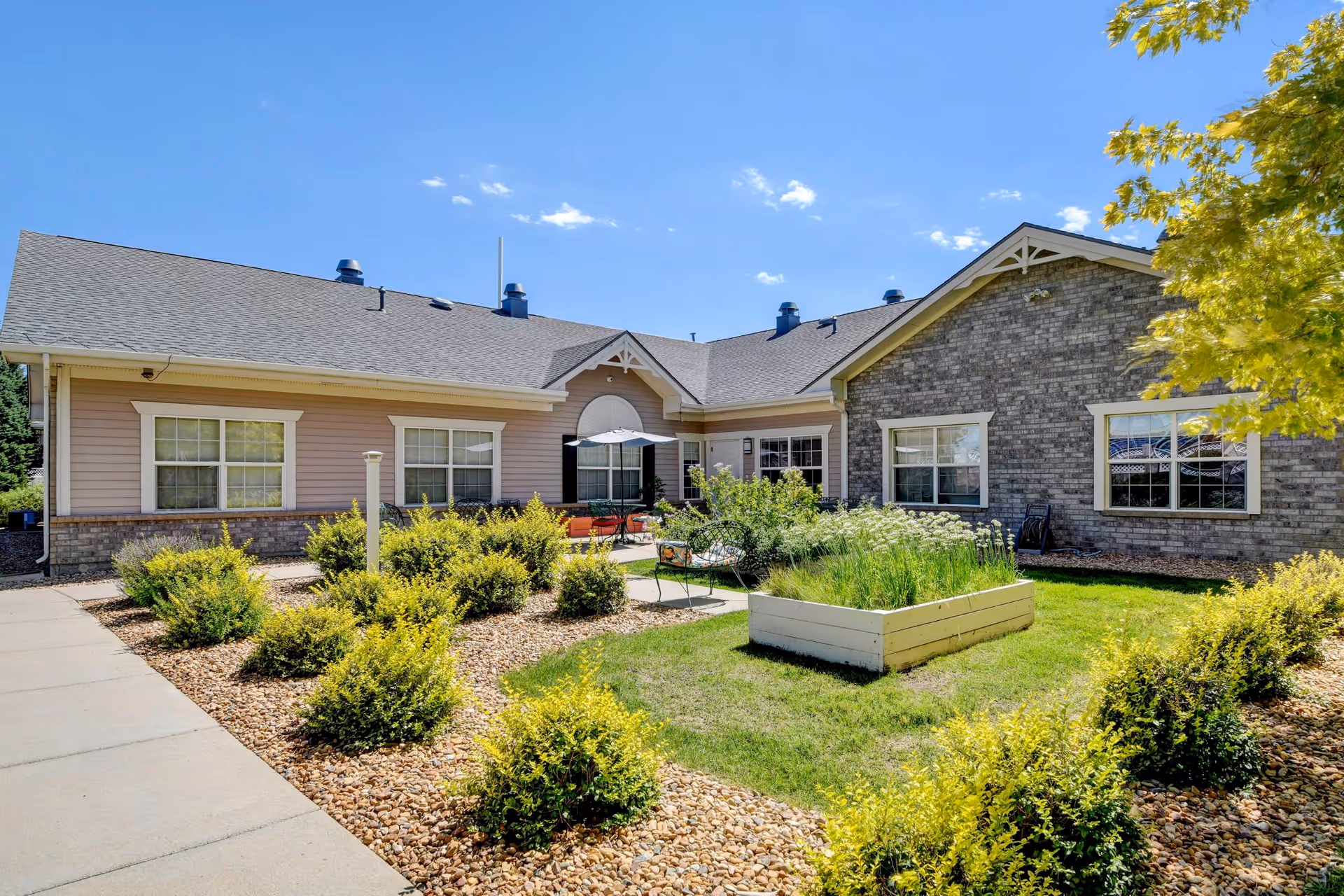 Outdoor courtyard area of a senior living facility with a paved walkway, green bushes, a raised garden bed, and patio furniture including a table with an umbrella. The building has a combination of brick and siding exterior with multiple windows under a clear blue sky.