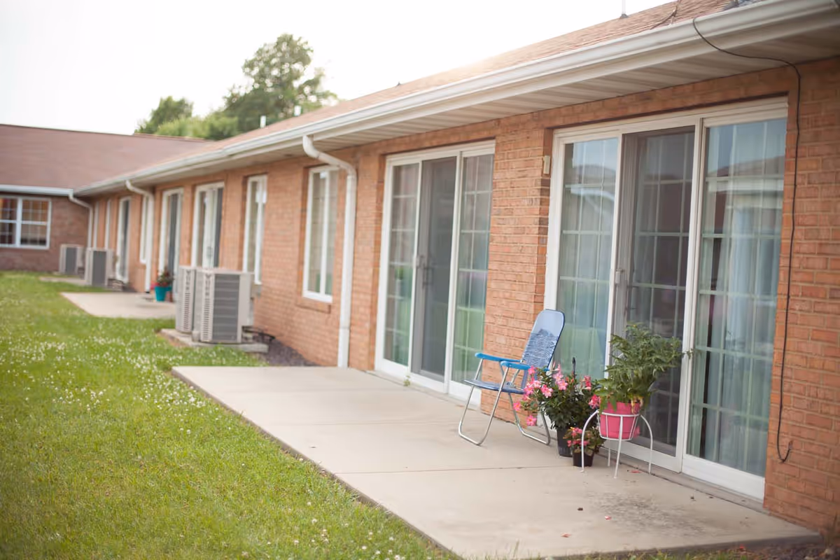 Brick single-story building exterior showing sliding glass patio doors, a concrete walkway, a blue chair and potted flowers.
