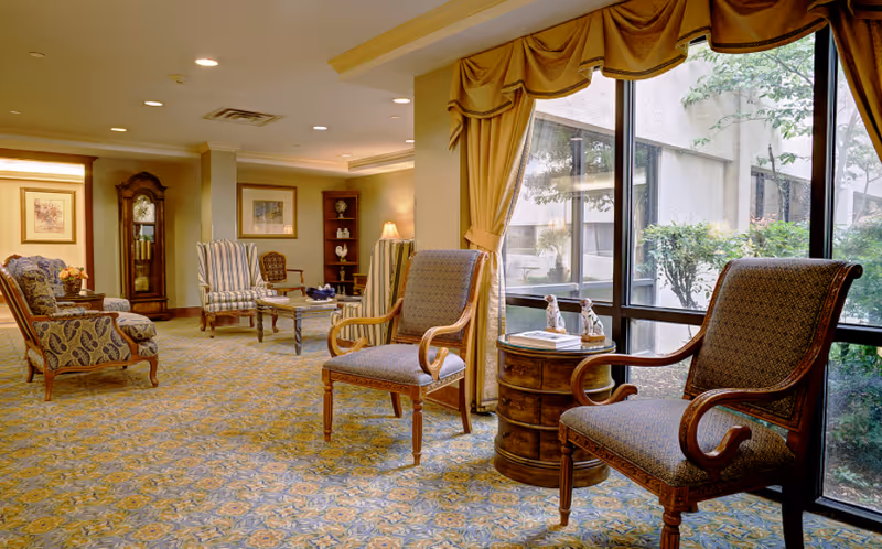 A cozy senior living facility common area with patterned carpet, several upholstered chairs, a wooden side table with decorative items, large windows with yellow curtains, and a grandfather clock in the background.