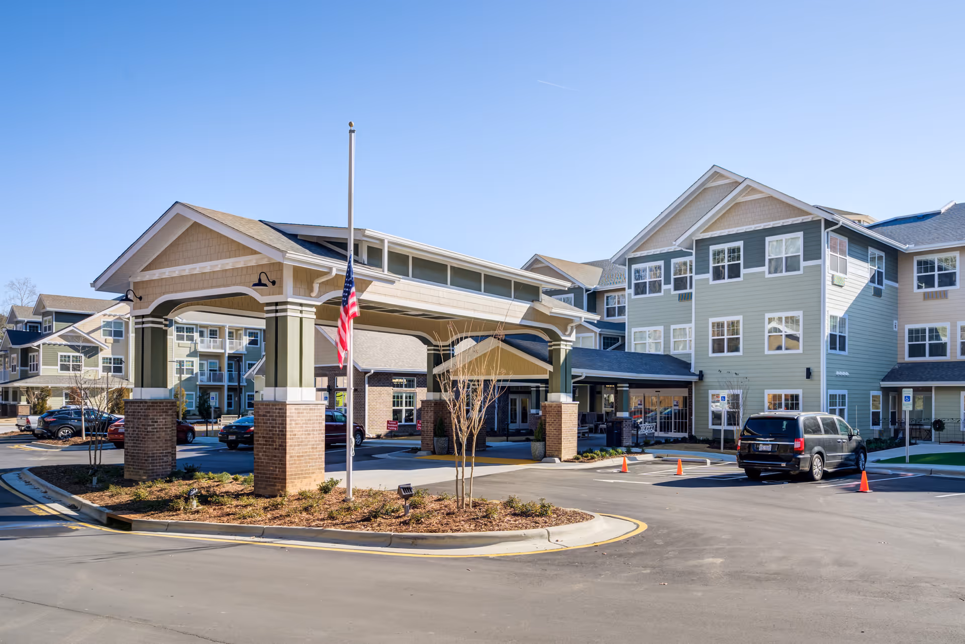 Exterior view of Azalea Estates Gracious Retirement Living facility showing a covered entrance with brick pillars, an American flag on a flagpole, multiple parked cars, and a multi-story building with many windows under a clear blue sky.