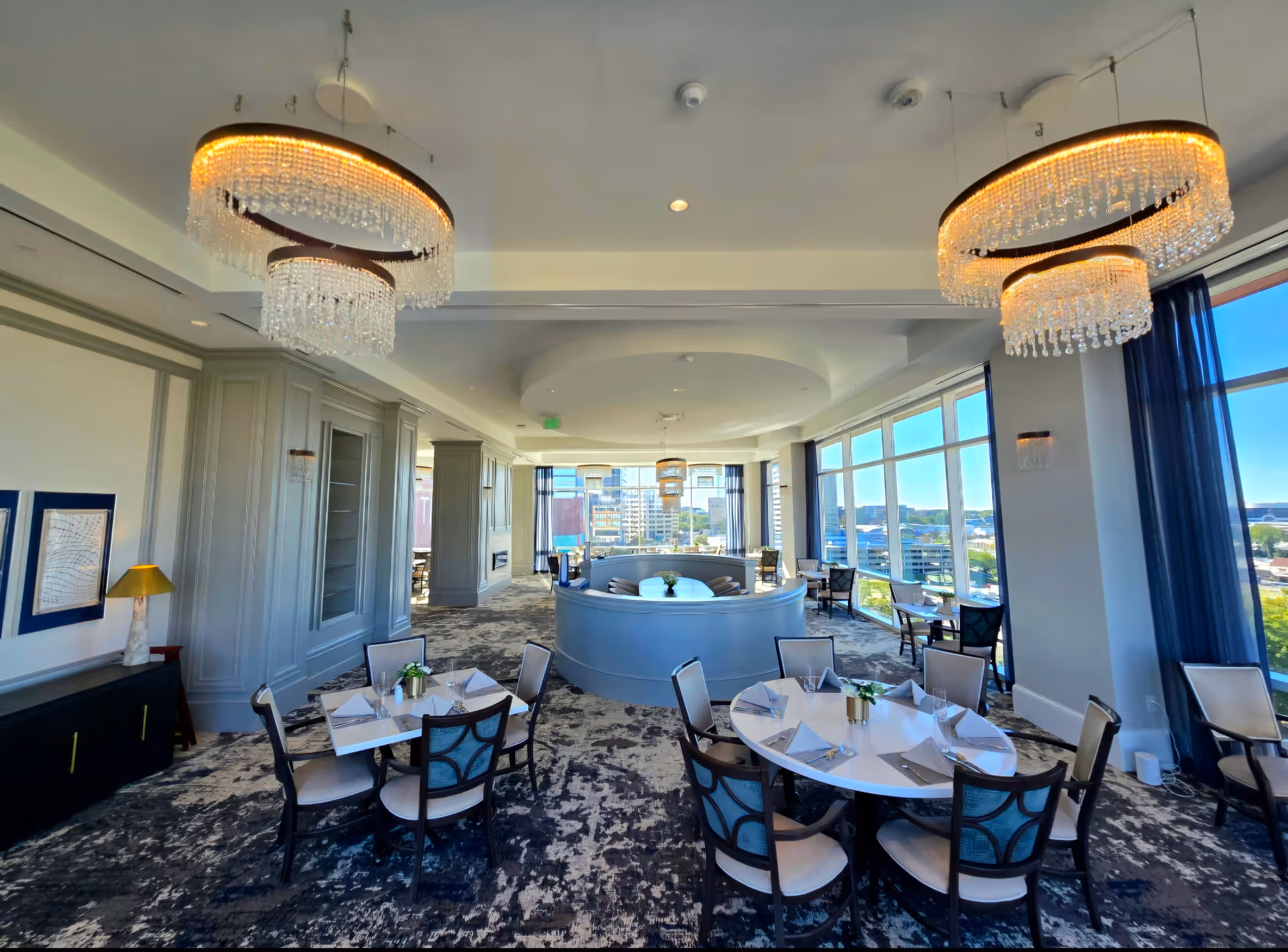 A bright and elegant dining room with round tables set with white napkins and silverware. The room features large windows with blue curtains allowing natural light and city views. There are modern chandeliers with crystal details hanging from the ceiling, and a curved booth seating area in the center. The carpet has a patterned design, and the walls are painted in light colors with decorative molding.