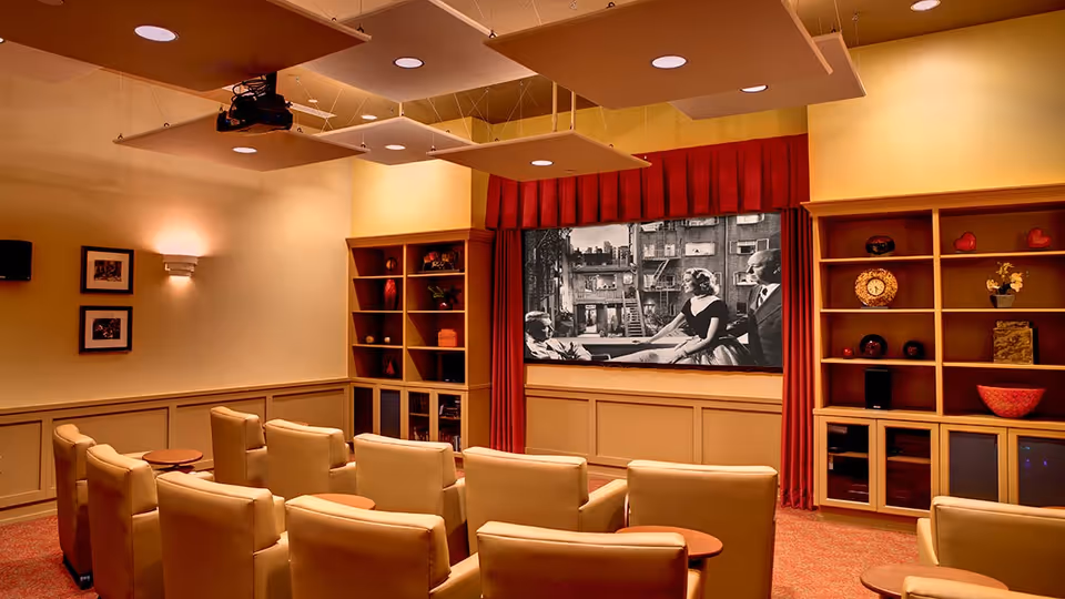 A cozy home theater room with beige leather recliner chairs arranged in rows facing a large screen showing a black and white movie. The room has warm lighting, built-in wooden shelves with decorative items, and red curtains framing the screen.