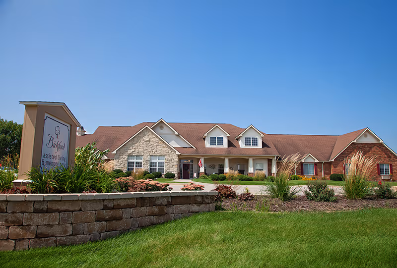 Exterior view of a single-story senior living facility building with a mix of stone and brick facade, a brown roof, and a well-maintained lawn and landscaping under a clear blue sky. A sign in the foreground reads 'Bridford assisted living & memory care'.
