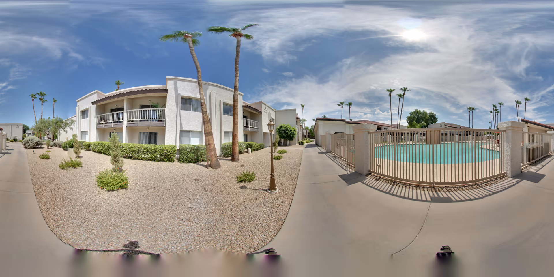Outdoor view of Pueblo Norte Senior Living facility showing a white two-story building with balconies, surrounded by desert landscaping with gravel and palm trees. To the right, there is a fenced swimming pool under a partly cloudy sky.